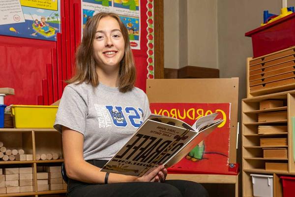 A smiling Indiana State education student sits in an elementary classroom. She is wearing a grey shirt that says ISU in blue letters and has an image of Sycamore Sam’s head inside the U. She is holding a book with the title “Giraffe Problems” that has the image of a giraffe’s neck on it. The name of the author, Jory John, and the illustrator, Lane Smith, are also visible at the top of the book. Behind the student is a red sign with the word “Corduroy” on it.