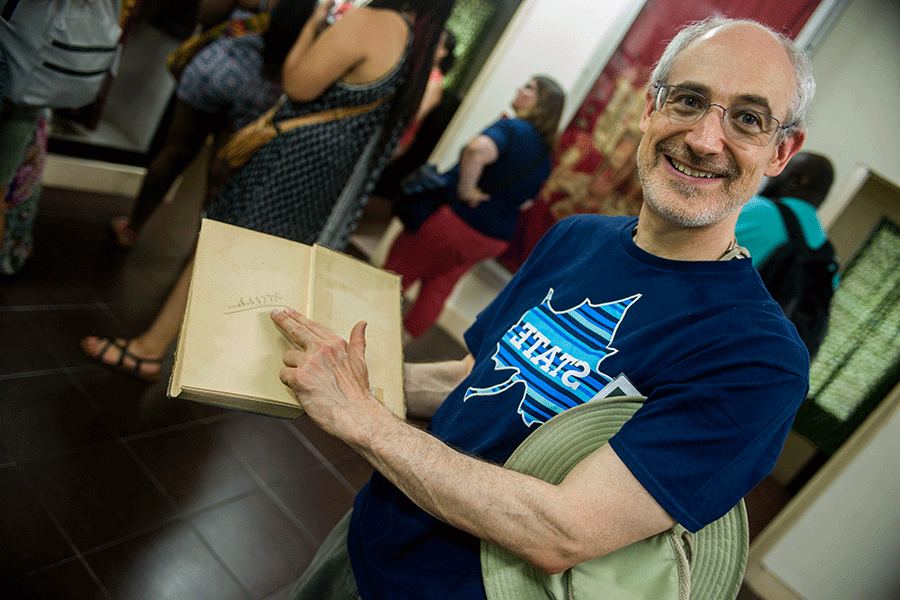 Older male adult in navy Indiana State University t-shirt smiles while pointing to a signature written in an old book.