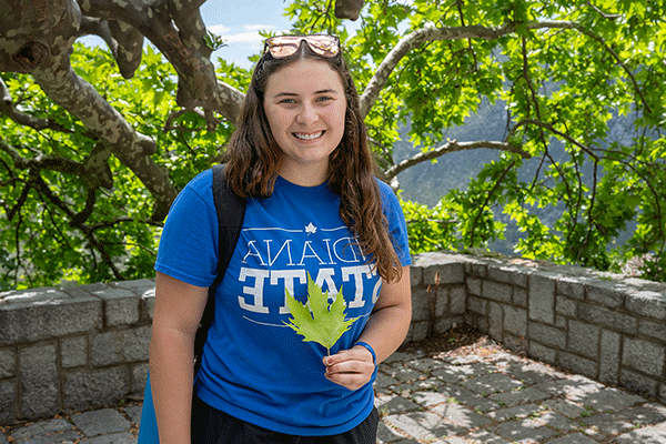 A female student with long brown hair poses outside with sunglasses on top of her head. She wears a blue Indiana State T-shirt and a backpack. She holds a green leaf. Behind her is a row of brick stones and tree branches.