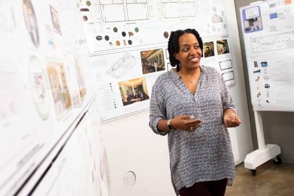 Azizi Arrington-Slocum, a Black female professor of interior architecture design, stands before three walls of interior architecture blueprints and plans. She has shoulder-length black hair, and she wears a grey shirt and black pants.