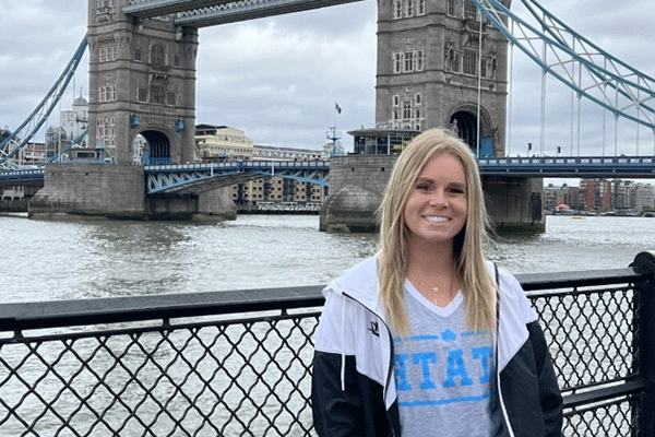 A white female student with straight blond hair poses in front of the London Bridge in London, England. She wears a light grey T-shirt with STATE in blue lettering and a black-and-white rain jacket. A river is visible behind her.